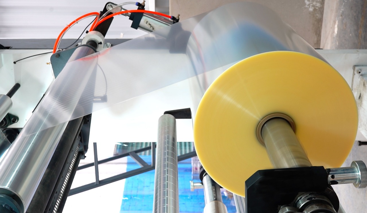 Bobina de material pl&aacute;stico transparente y sin imprimir, en pleno proceso de desenrollado o conversi&oacute;n dentro de una l&iacute;nea de producci&oacute;n industrial.ch